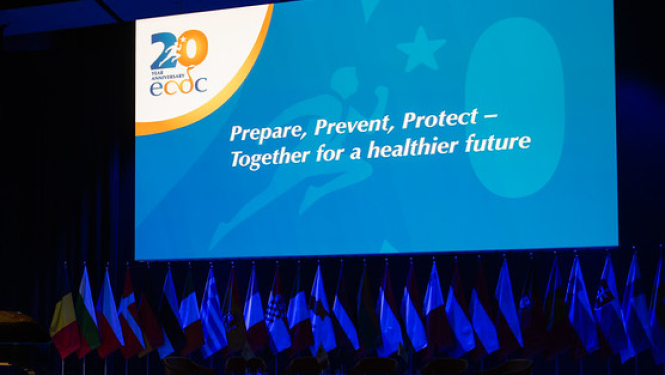 Eiropas slimību profilakses un kontroles centra 20 gadu svinību oficiālais foto. Svinību zāle ar plakātu. Uzraksts uz plakāta: "Gatavoties, novērsts, pasargāt".