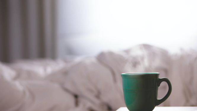 gaiša istaba ar aizkariem un centrā balta gultasveļa, blakus galds uz kura stāv zaļa krūze