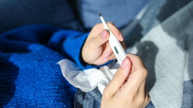 Sieviete gultā, zilā džemperī tur rokā termometru