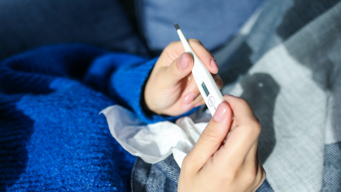 cilvēks zilā džemperī guļ gultā un rokās termometrs