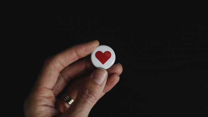 uz melns fona cilvēka roka tur baltu, apaļu piespraudi ar sirds attēlu