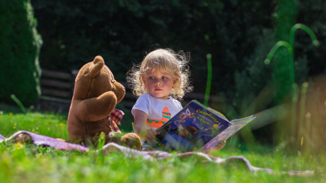 Maza meitene ar grāmatu un spēļu lāci sēž zaļā zālē