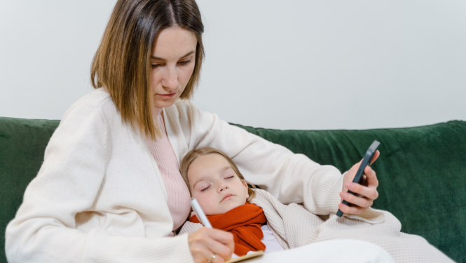 Sieviete sēž dīvānā ar mazuli klēpī un mēra bērnam temperatūru