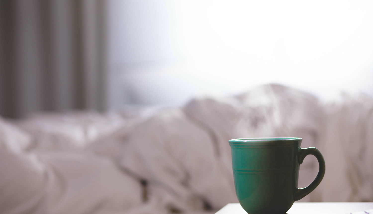 gaiša istaba ar aizkariem un centrā balta gultasveļa, blakus galds uz kura stāv zaļa krūze