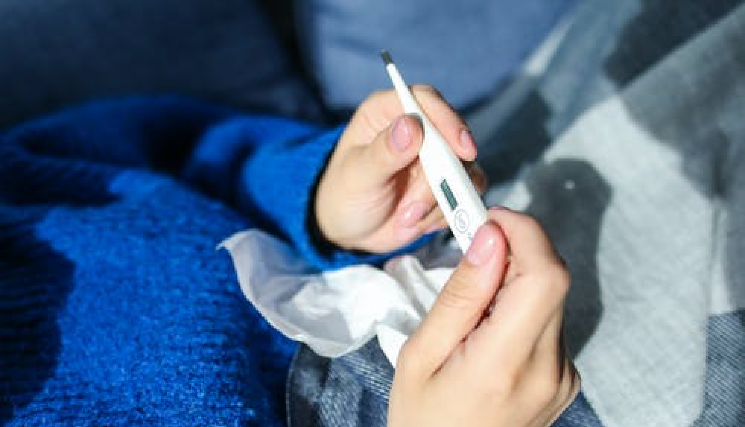 Sieviete gultā, zilā džemperī tur rokā termometru