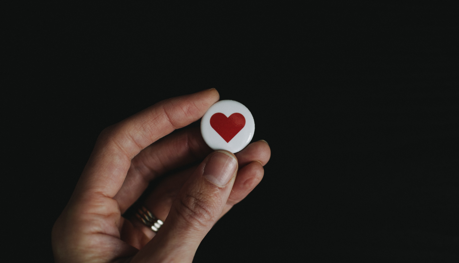 uz melns fona cilvēka roka tur baltu, apaļu piespraudi ar sirds attēlu