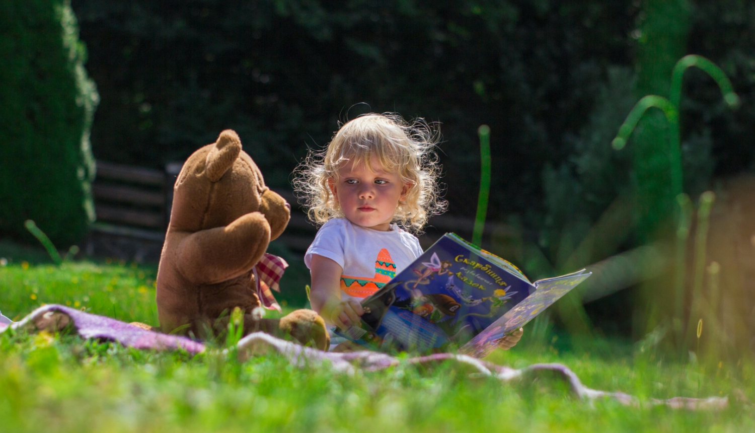 Maza meitene ar grāmatu un spēļu lāci sēž zaļā zālē