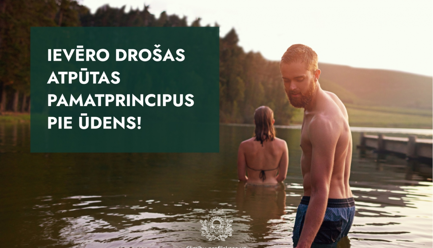 sieviete stāv ezerā ar muguru un vīrietis zilās peldbiksēs pagriezies ar seju