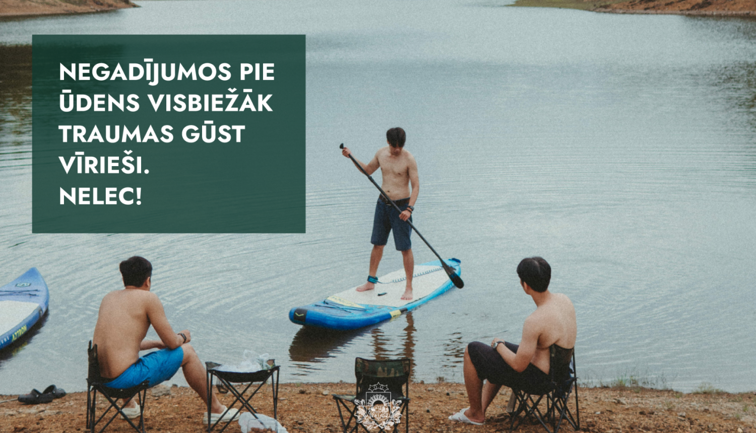 cilvēki atpūšas pie ūdens, divi sēž krastā, viens uz supa ūdenī