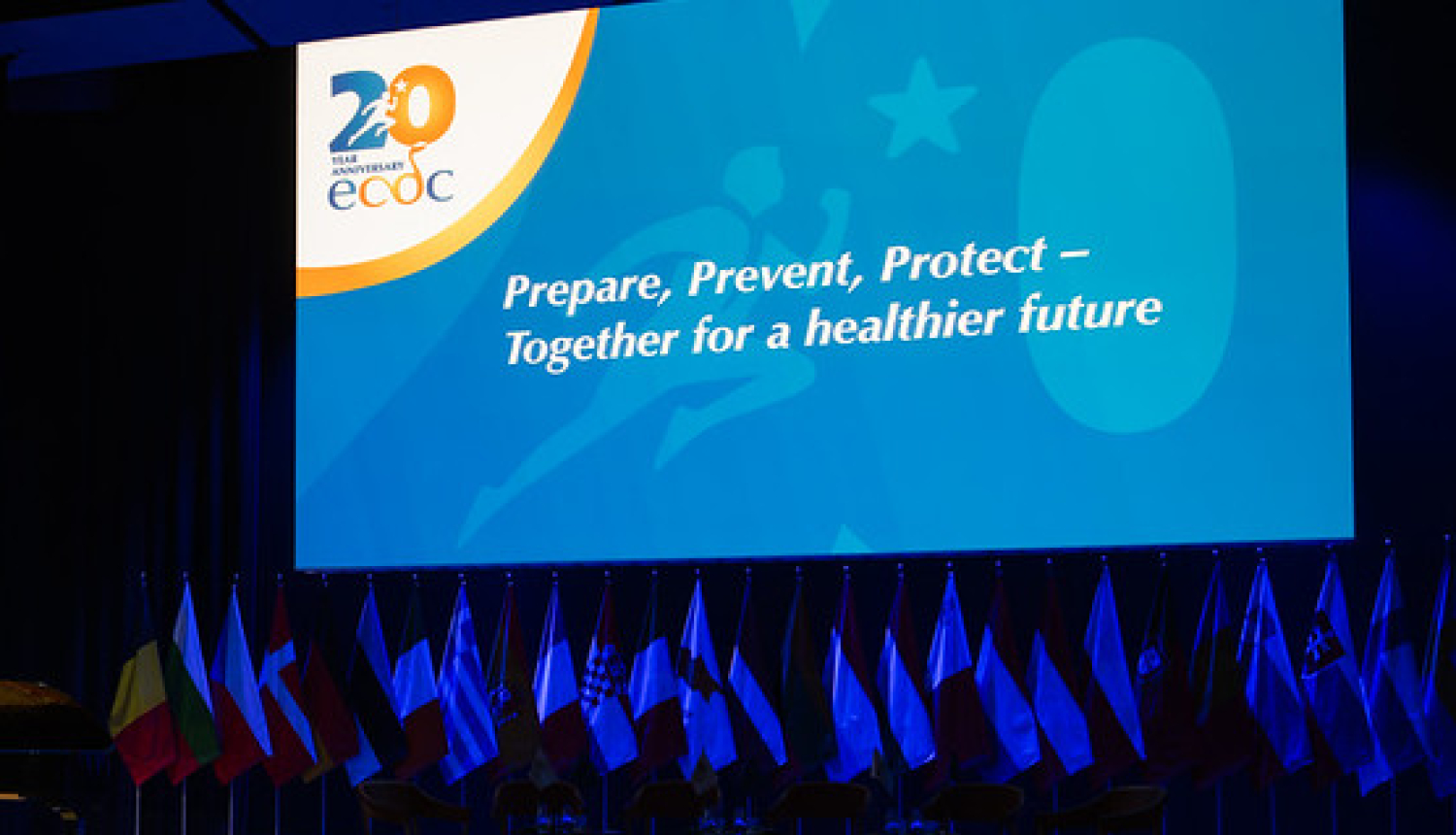Eiropas slimību profilakses un kontroles centra 20 gadu svinību oficiālais foto. Svinību zāle ar plakātu. Uzraksts uz plakāta: "Gatavoties, novērsts, pasargāt".